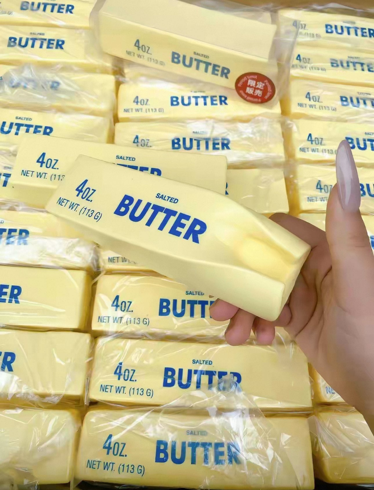 A hand holding a hachimi butter squishy toy with 4 oz stick of salted butter in front of a stacked background of similar butter packages.
