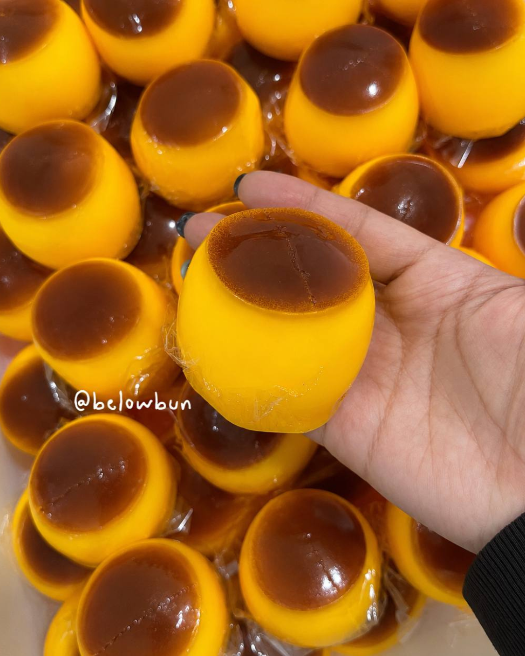 A hand holds a small yellow caramel pudding scrunchy squishy toy dessert with a glossy caramelized brown top, surrounded by many similar desserts in a tray