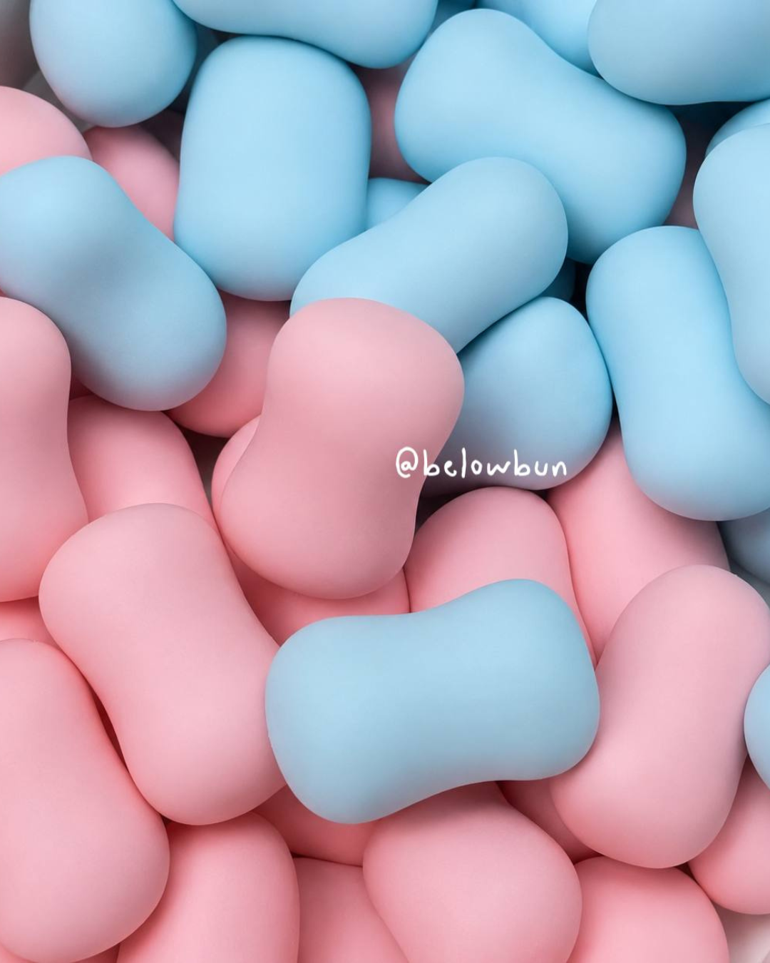 A close-up of smooth, rounded candy-like slushy squishy toy pieces in pastel pink and light blue, filling the frame. A small white '@belowbun' watermark appears near the center.