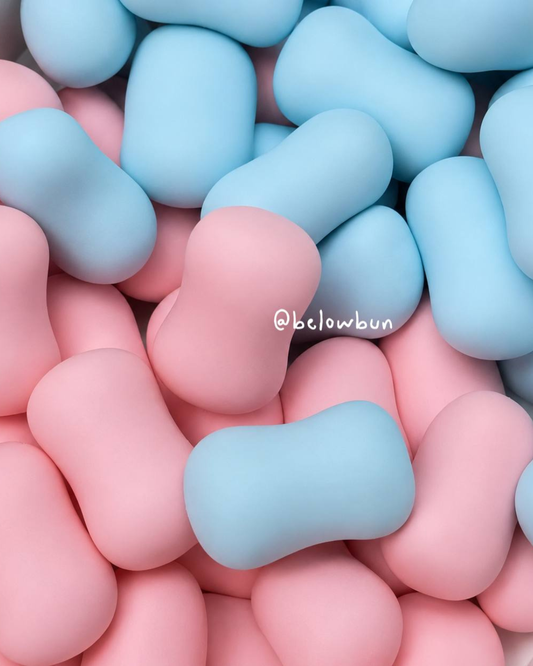 A close-up of smooth, rounded candy-like slushy squishy toy pieces in pastel pink and light blue, filling the frame. A small white '@belowbun' watermark appears near the center.