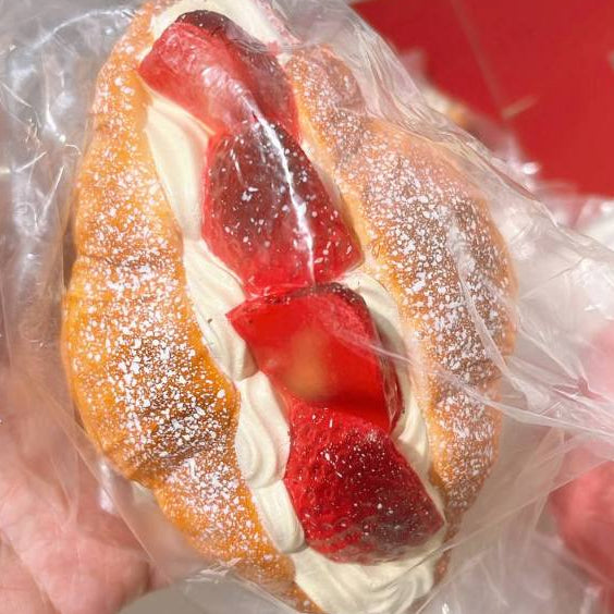 A close-up of a strawberry-filled croissant squishy toy, wrapped in clear plastic and held in a hand. The pastry is dusted with powdered sugar and filled with white cream and red strawberry pieces.